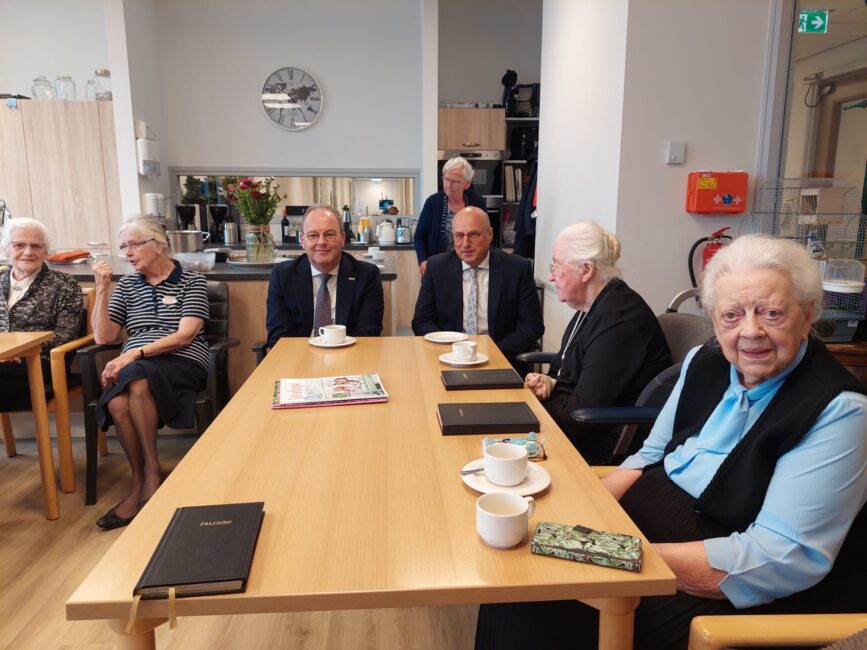 Een groep mensen zit aan een tafel in een gemeenschappelijke ruimte, met koffie en documenten voor zich op tafel.