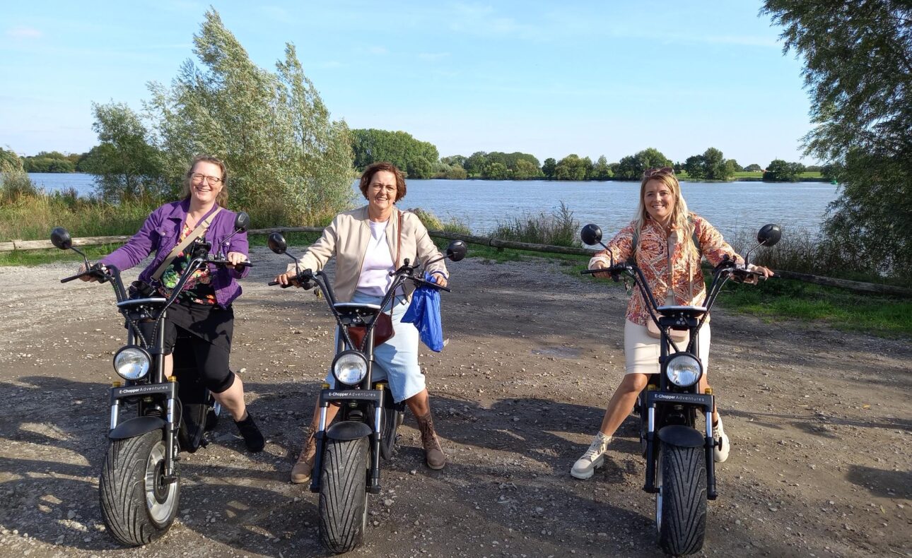 Drie mensen poseren op elektrische scooters bij een meer, omringd door bomen, op een zonnige dag met een blauwe lucht.