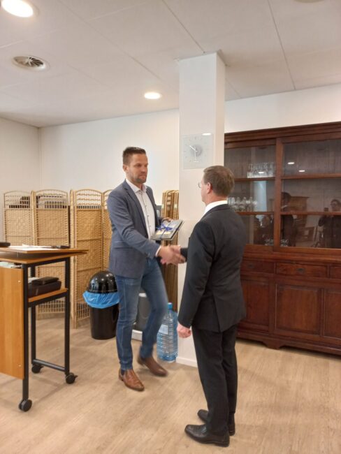 Twee mannen schudden elkaar de hand in een kantoorruimte. Een man draagt een blauw pak, de ander een zwart pak.