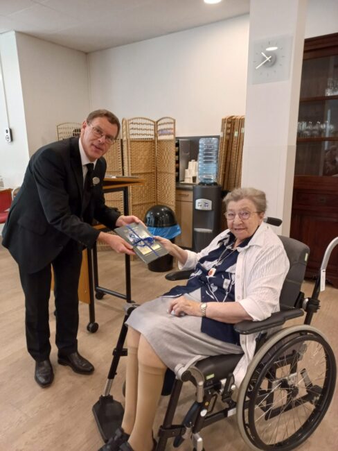 Een man in een pak overhandigt een boek aan een oudere vrouw in een rolstoel in een kamer met opgestapelde stoelen.
