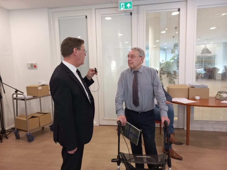 Twee mannen in gesprek in een kantooromgeving. De man rechts gebruikt een rollator. Op de achtergrond staan dozen.