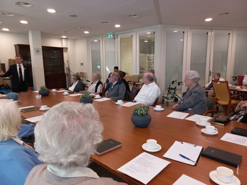 Een groep ouderen zit rond een vergadertafel in een lichte ruimte. Er liggen notitieblokken en kopjes koffie op tafel.