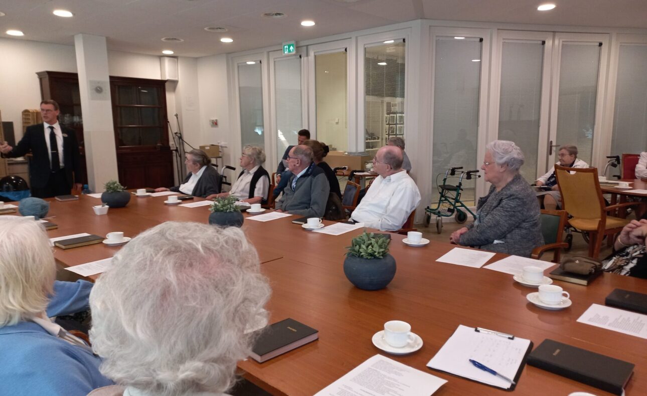 Een groep ouderen zit rond een vergadertafel in een lichte ruimte. Er liggen notitieblokken en kopjes koffie op tafel.