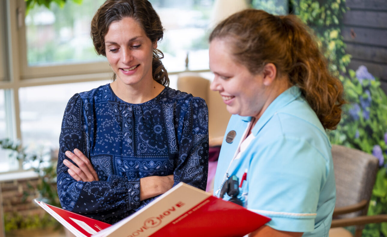 Twee vrouwen bekijken samen een rode map in een lichte kamer. De vrouw rechts draagt een lichtblauw uniform en glimlacht.
