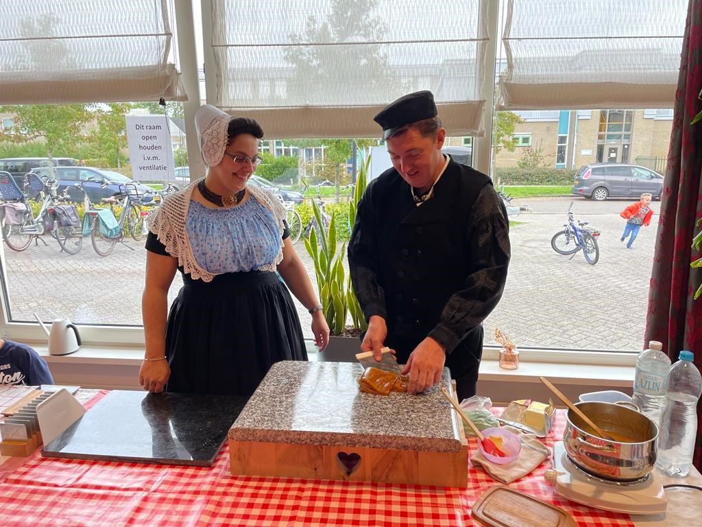 Twee mensen in traditionele Nederlandse klederdracht bereiden eten voor op een tafel met een rood-wit geruit tafelkleed.
