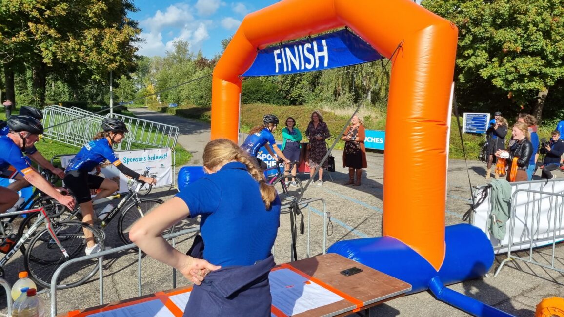 Fietsers in blauwe shirts passeren de finishlijn van een wielerwedstrijd. Een vrouw kijkt naar de deelnemers bij de finish.