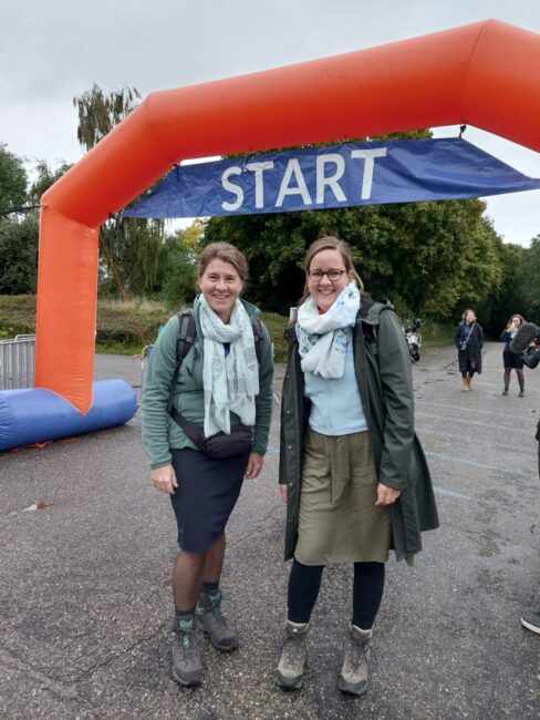 Twee vrouwen staan onder een oranje startboog, klaar voor een sportevenement. Ze dragen wandelschoenen en sjaals.