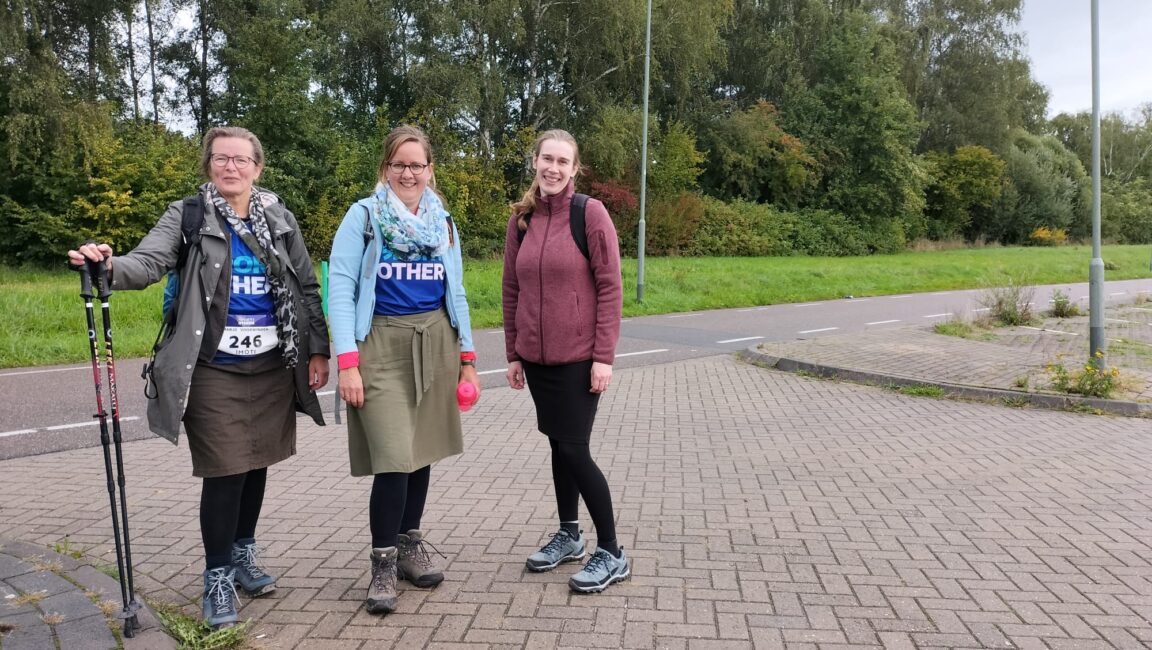 Drie vrouwen staan buiten op een trottoir, gekleed in sportieve kleding. Twee dragen blauwe shirts met de tekst "FOR OTHERS".