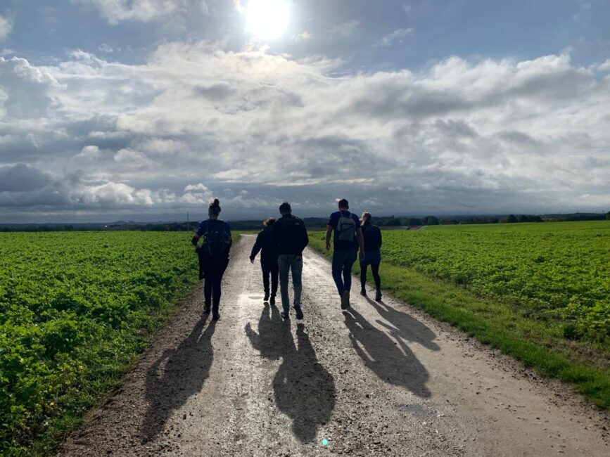 Vijf mensen wandelen op een landweg tussen groene velden onder een bewolkte hemel met zonnestralen die door de wolken breken.