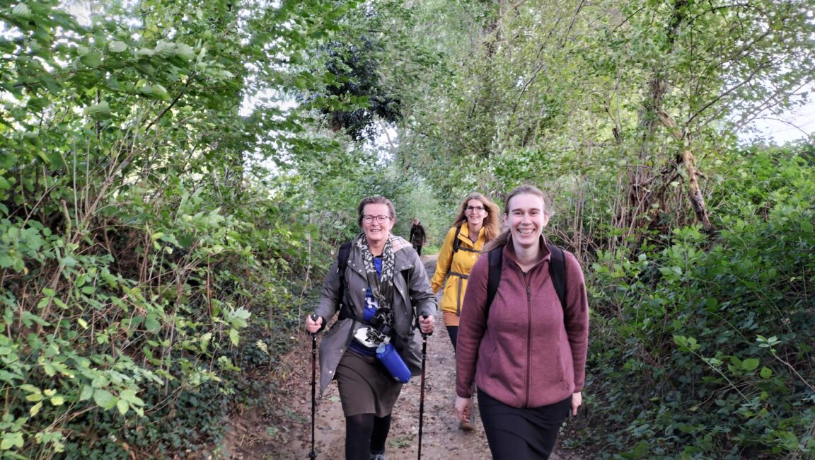 Drie lachende mensen wandelen op een bosachtig pad, omringd door groene bomen en struiken, met wandelstokken en rugzakken.