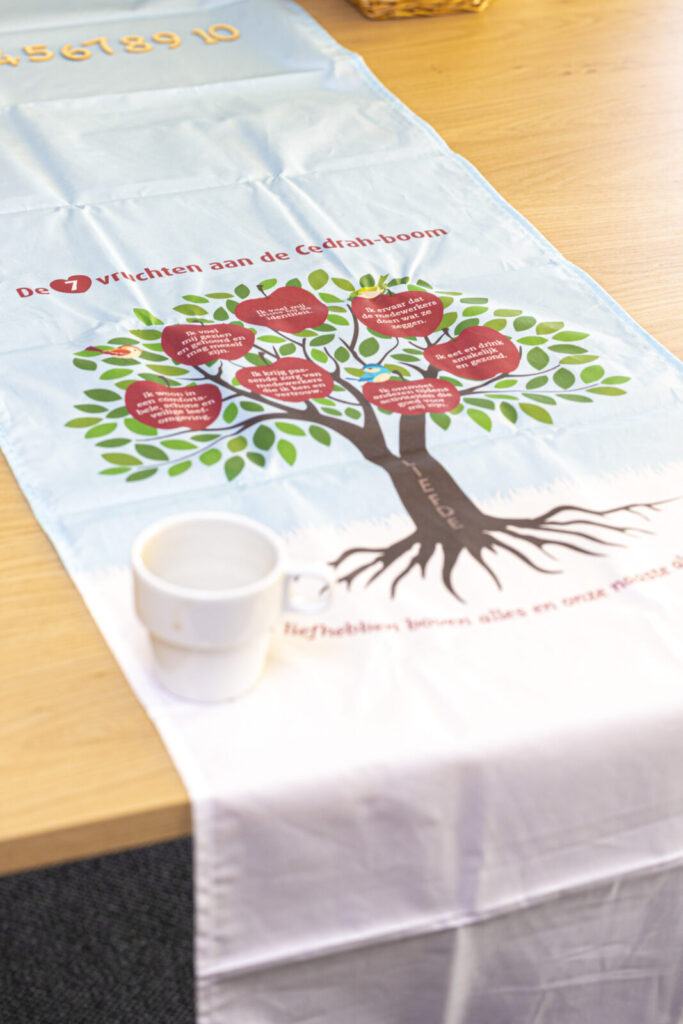 Een tafelkleed met een boomillustratie en tekst, ligt op een houten tafel. Er staat een witte mok op het kleed.