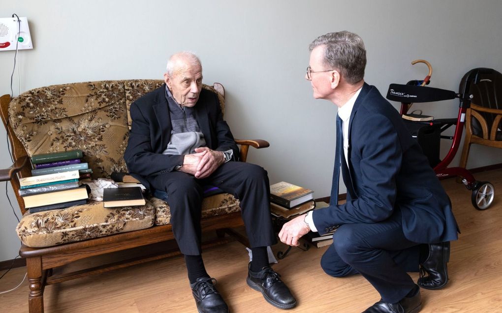 Twee mannen in gesprek; een oudere man zit op een bank met boeken naast zich, terwijl een andere man voor hem knielt.