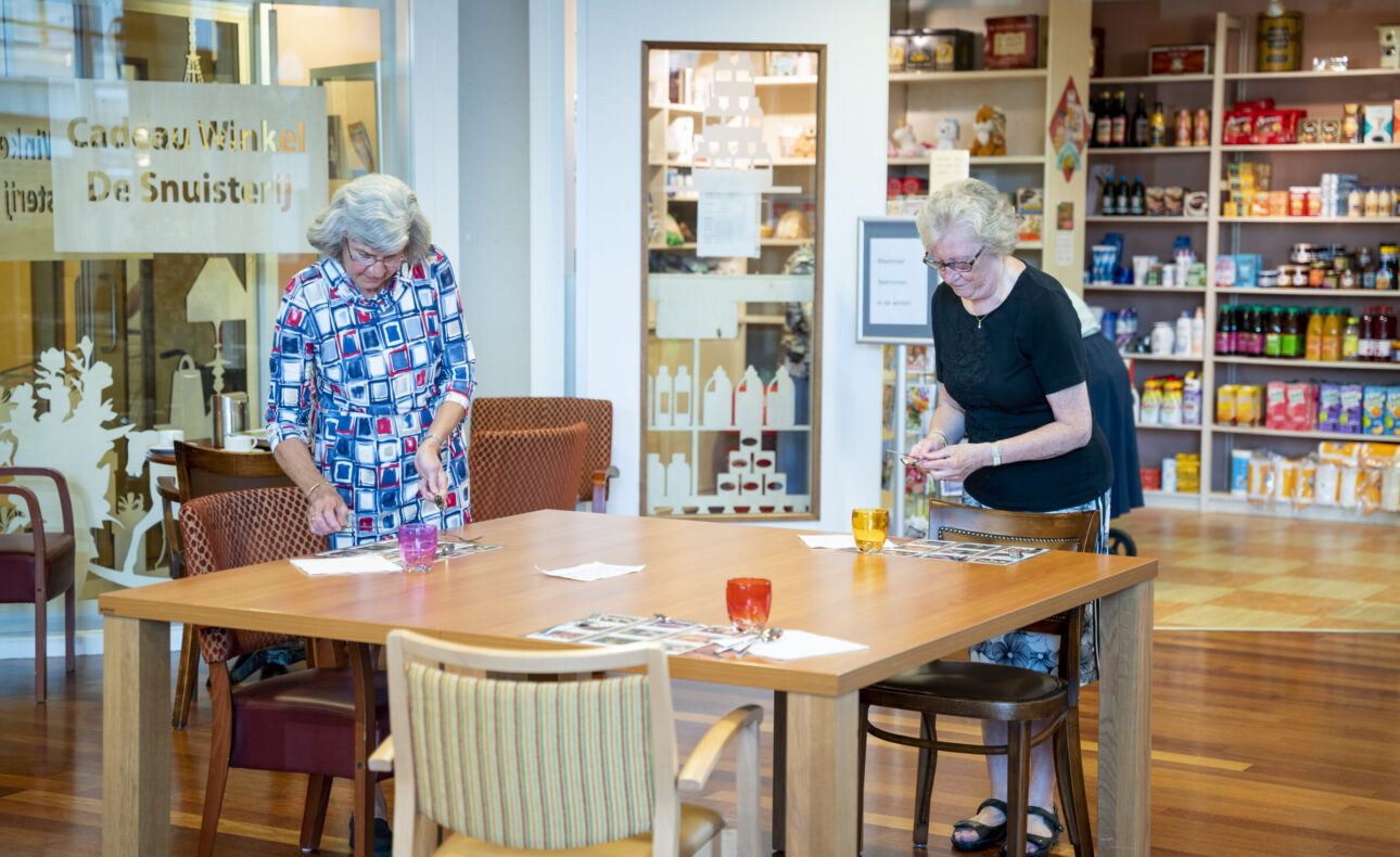 Twee oudere vrouwen spelen een spel aan een houten tafel in een gemeenschappelijke ruimte met een winkel op de achtergrond.