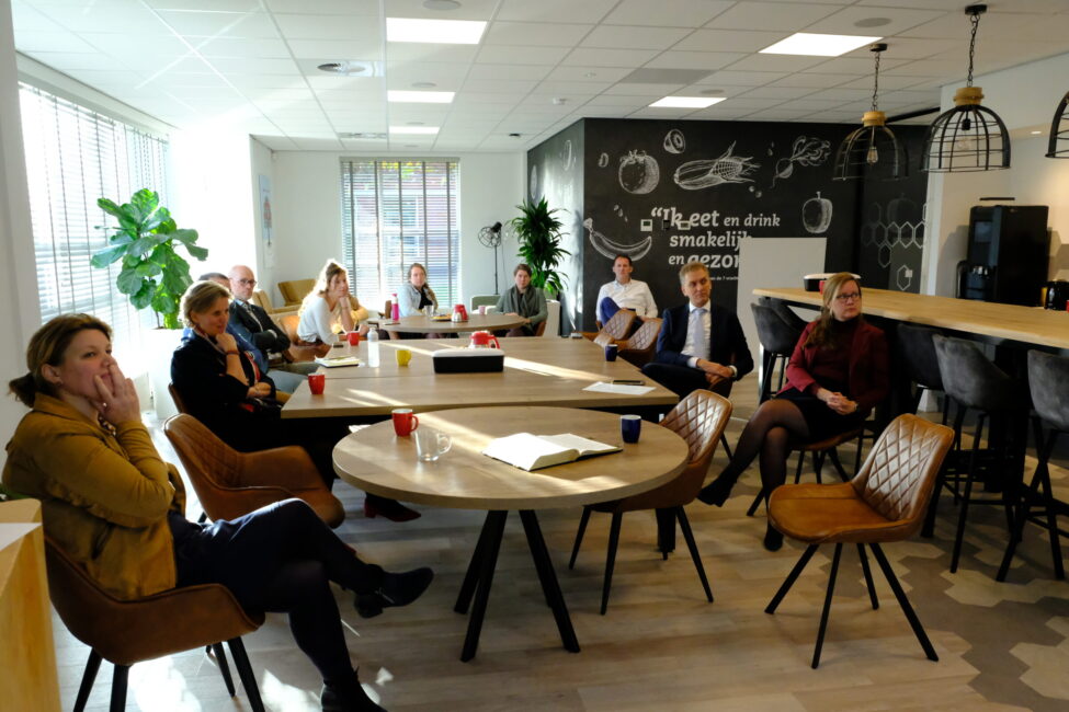 Groep mensen zit in een moderne vergaderruimte met ronde tafels en een krijtbordmuur. Ze luisteren aandachtig naar een spreker.