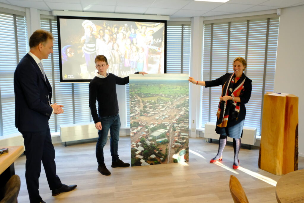 Drie mensen staan in een kamer en houden een grote foto vast. Op de achtergrond is een scherm met een kleurrijke afbeelding.