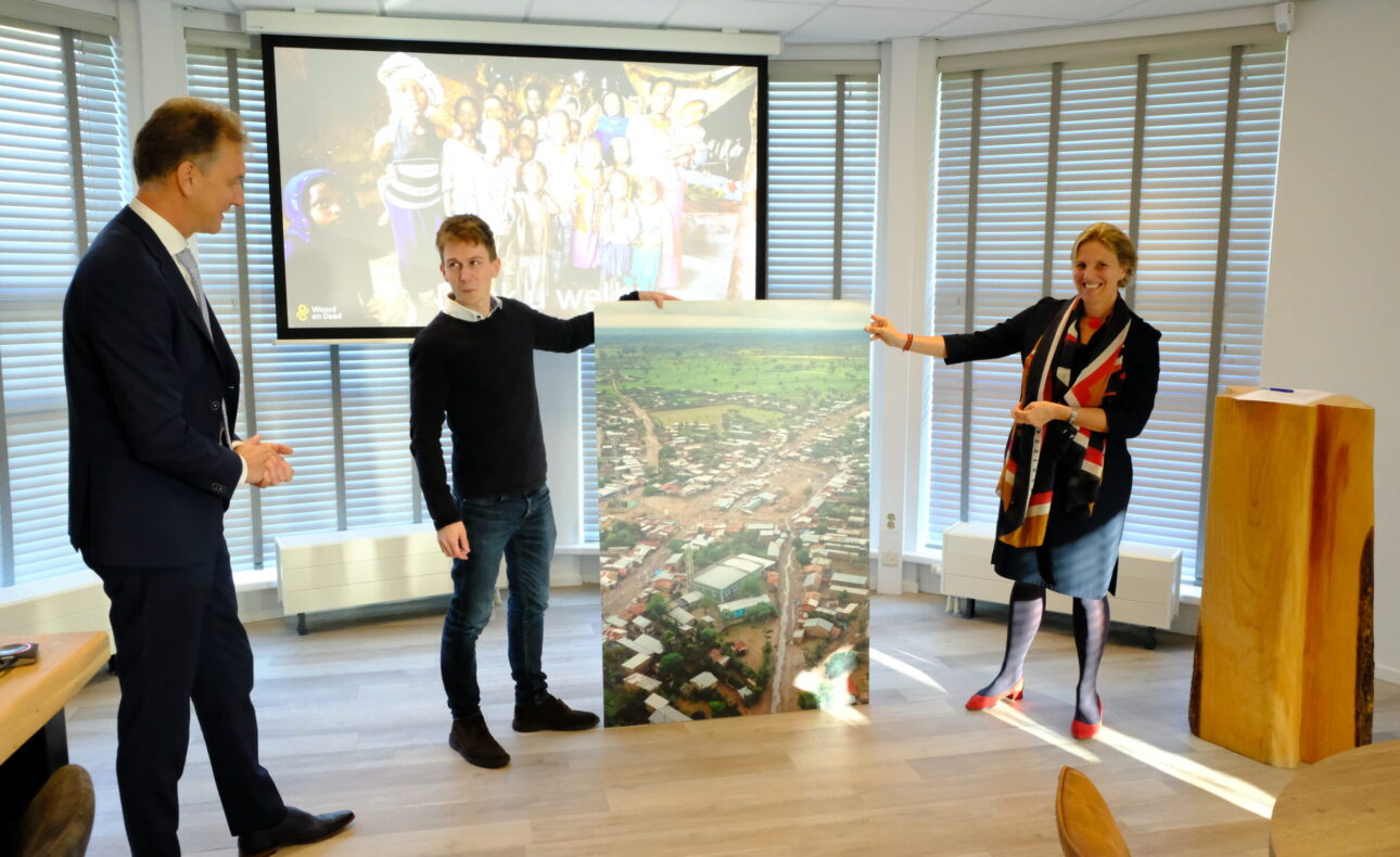 Drie mensen staan in een kamer en houden een grote foto vast. Op de achtergrond is een scherm met een kleurrijke afbeelding.