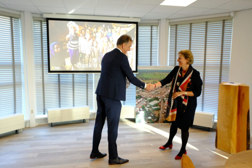 Twee mensen schudden elkaar de hand in een kantoorruimte met een projectiescherm en een groot schilderij op de achtergrond.