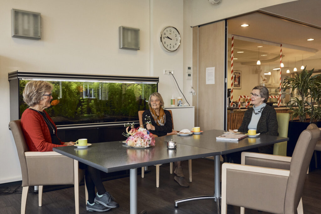 Drie vrouwen zitten aan een tafel in een gezellige ruimte met een groot aquarium op de achtergrond en drinken koffie.