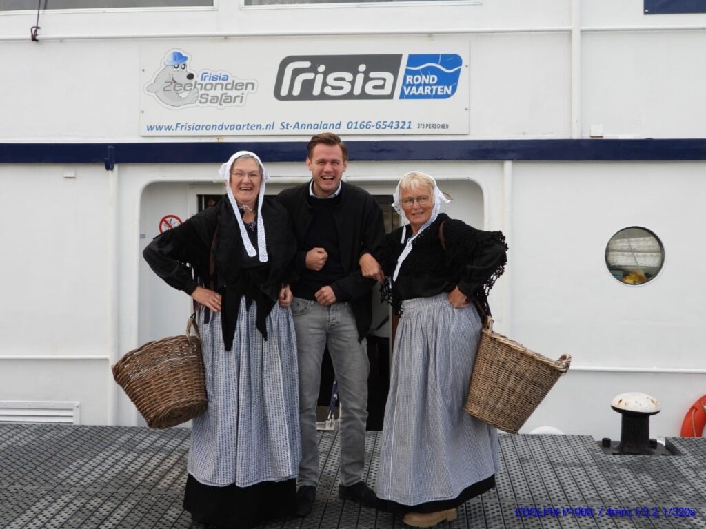 Een man poseert tussen twee vrouwen in traditionele Nederlandse klederdracht met manden, voor een boot met Frisia-logo.
