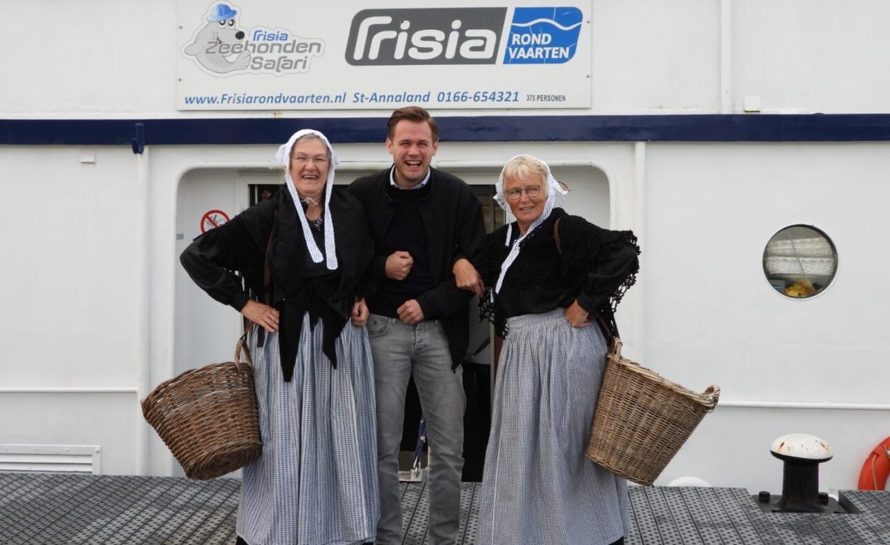 Een man poseert tussen twee vrouwen in traditionele Nederlandse klederdracht met manden, voor een boot met Frisia-logo.