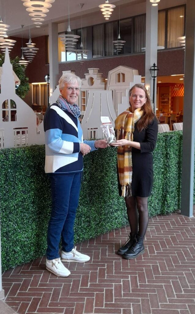 Twee mensen staan binnen en houden samen een boek vast. Op de achtergrond is een groene heg en een decoratief gebouw te zien.