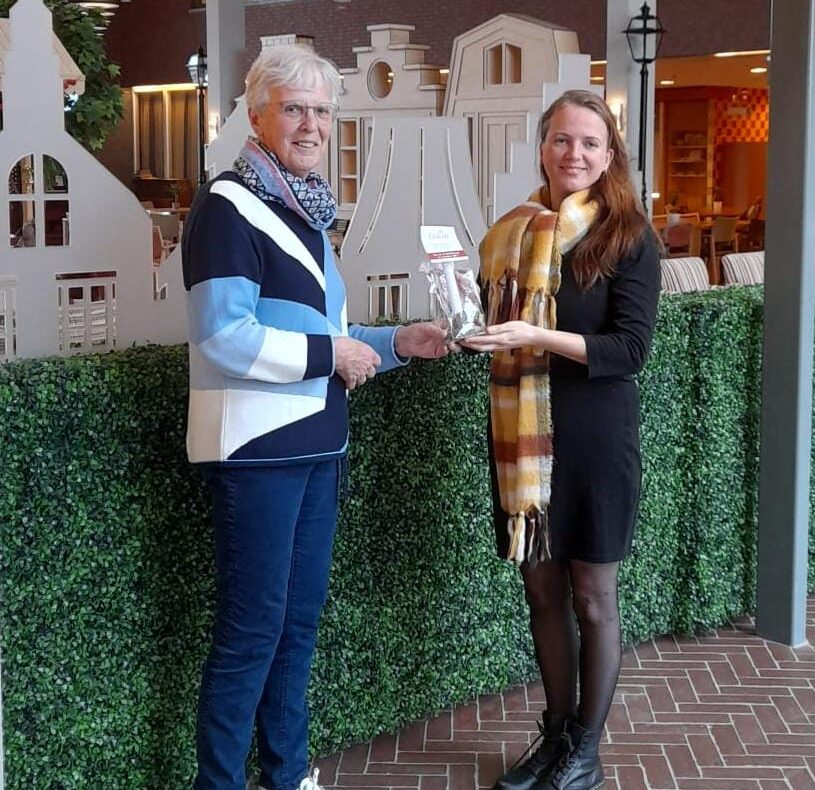 Twee mensen staan binnen en houden samen een boek vast. Op de achtergrond is een groene heg en een decoratief gebouw te zien.