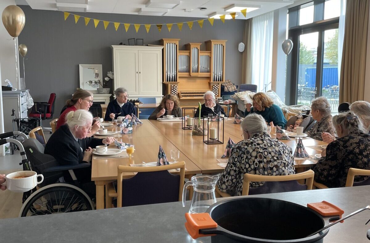 Een groep mensen zit rond een tafel in een gemeenschappelijke ruimte, versierd met gele vlaggetjes en ballonnen.