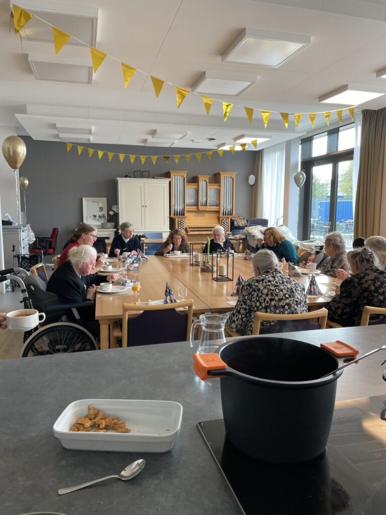 Een groep mensen zit rond een tafel in een gemeenschappelijke ruimte, versierd met gele vlaggetjes en ballonnen.
