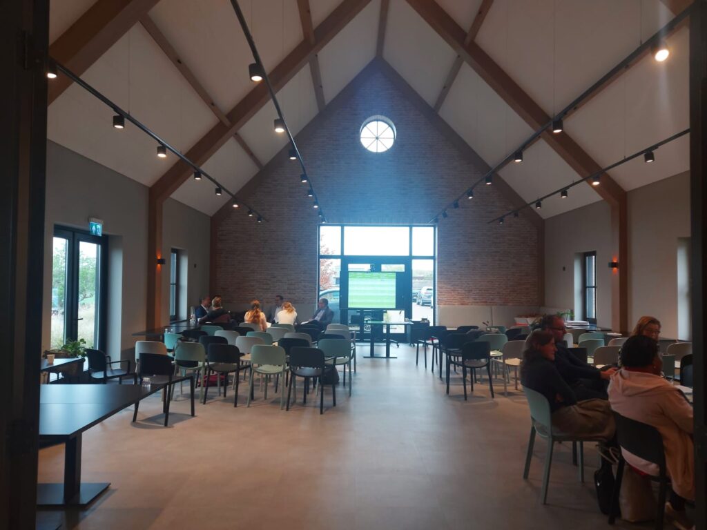 Een moderne, goed verlichte zaal met hoge plafonds, gevuld met mensen die aan tafels zitten. Grote ramen aan het einde.
