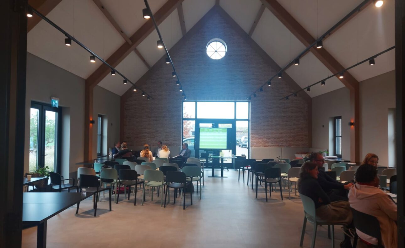 Een moderne, goed verlichte zaal met hoge plafonds, gevuld met mensen die aan tafels zitten. Grote ramen aan het einde.