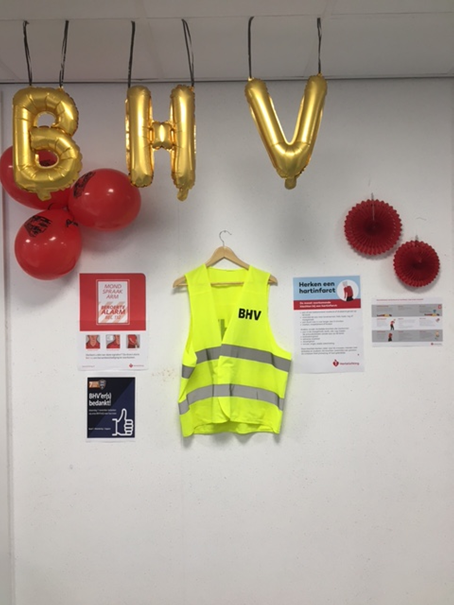 Gele BHV-veiligheidsvest hangt aan muur, omringd door rode ballonnen en posters. Gouden BHV-ballonnen bovenaan.