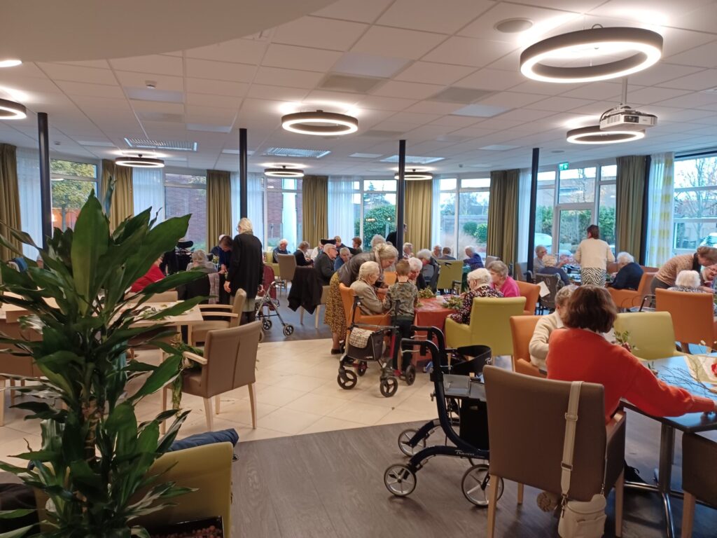 Een drukke gemeenschappelijke ruimte in een verzorgingstehuis, met ouderen die aan tafels zitten en met elkaar praten.