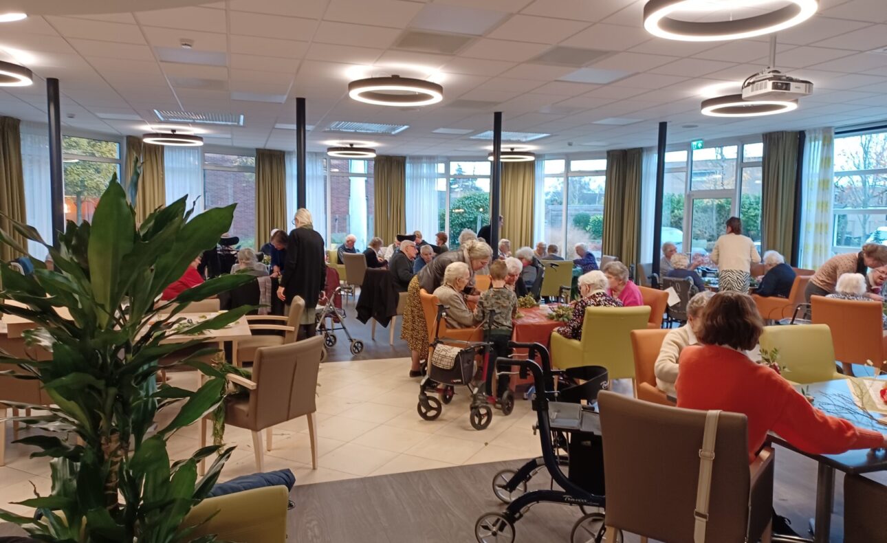 Een drukke gemeenschappelijke ruimte in een verzorgingstehuis, met ouderen die aan tafels zitten en met elkaar praten.