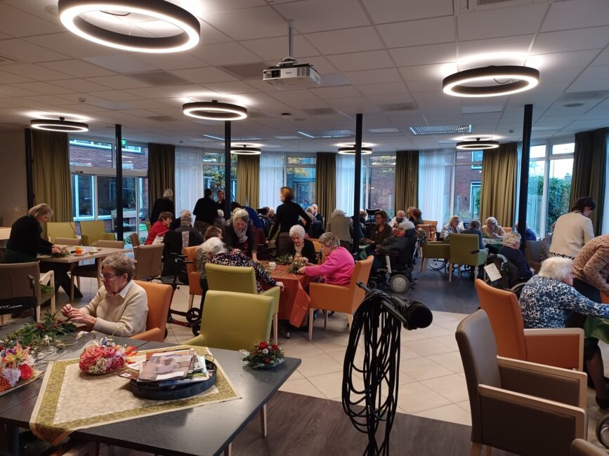 Groep ouderen zit in een gemeenschappelijke ruimte, bezig met knutselactiviteiten aan tafels. De sfeer is gezellig en ontspannen.