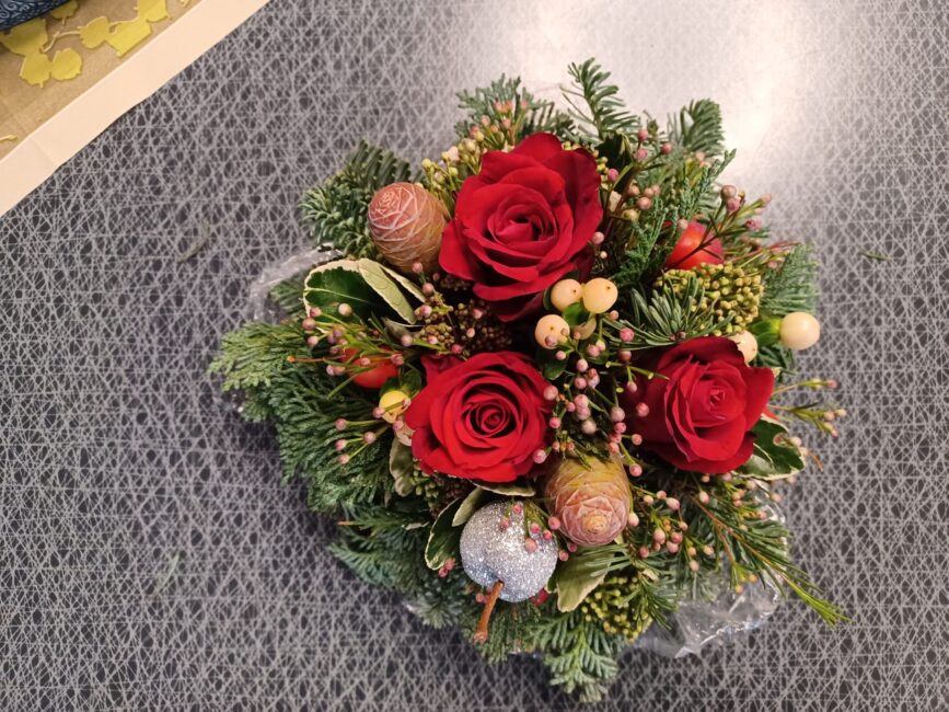 Een bloemstuk met rode rozen, groene takken en decoratieve bessen ligt op een grijs oppervlak. Het is feestelijk en kleurrijk.