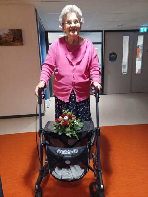 Oudere vrouw met grijs haar en roze vest staat met een rollator in een gang. Er ligt een boeket bloemen op de rollator.