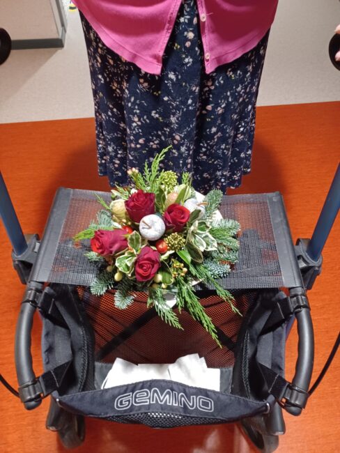 Een rollator met een bloemstuk van rode en witte bloemen en groen blad, geplaatst op de zitting. Een persoon staat erachter.