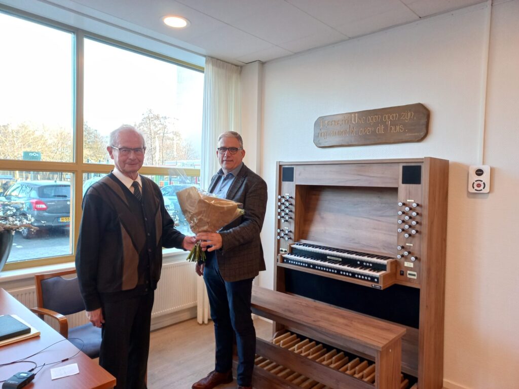 Twee mannen staan naast een orgel in een kamer. De man rechts houdt een boeket bloemen vast. Er is een raam op de achtergrond.