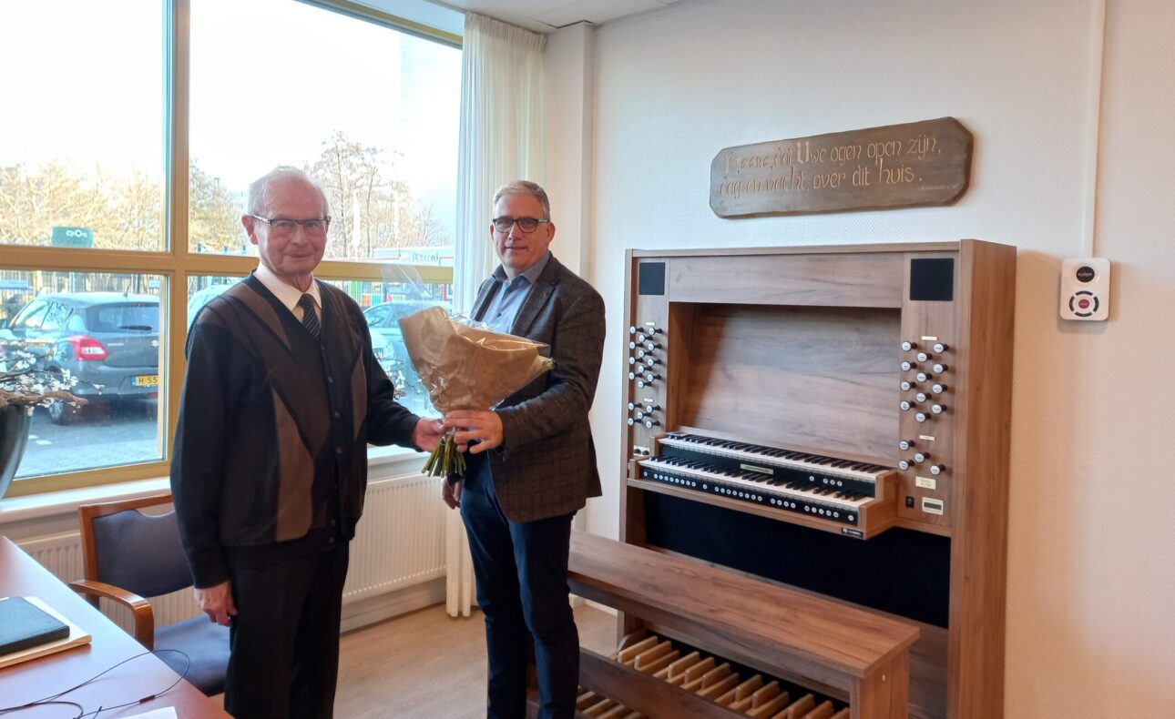 Twee mannen staan naast een orgel in een kamer. De man rechts houdt een boeket bloemen vast. Er is een raam op de achtergrond.