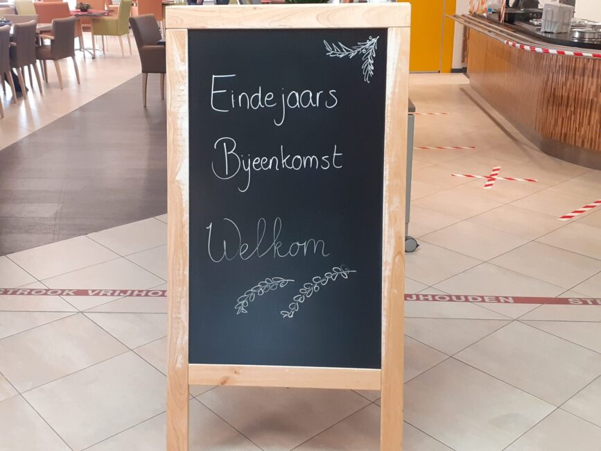 Zwarte krijtbord met de tekst "Eindejaars Bijeenkomst Welkom" in een lichte ruimte met tafels en stoelen op de achtergrond.