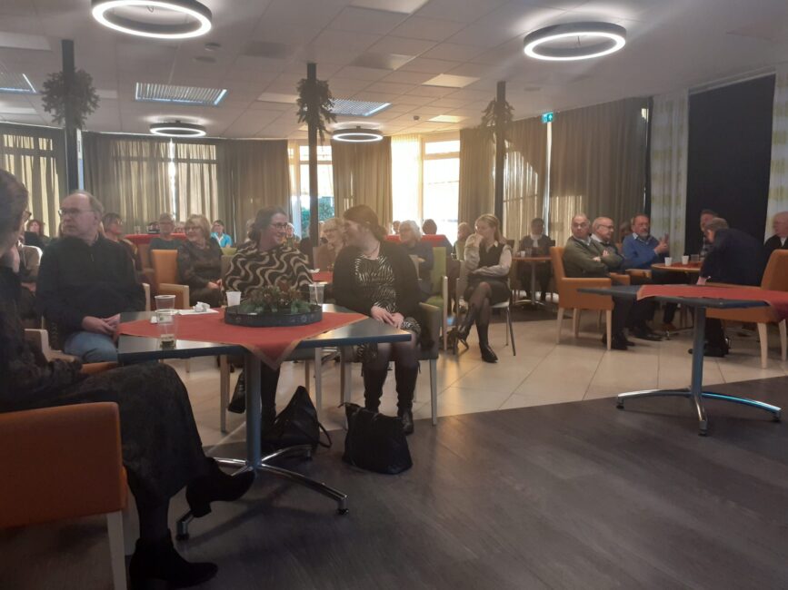 Groep mensen zit in een verlichte zaal met tafels en stoelen, bezig met een bijeenkomst of evenement. Tafels zijn gedekt.