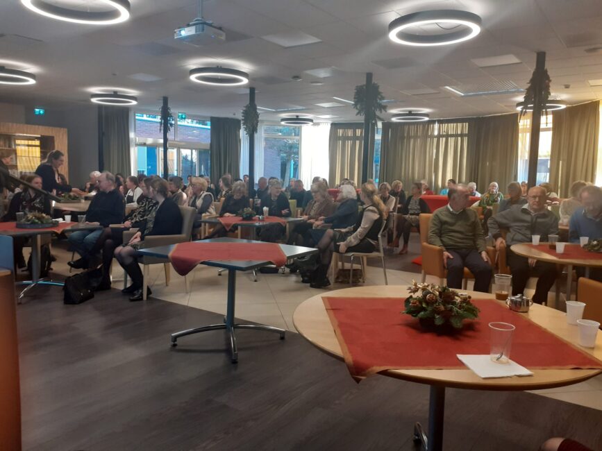 Een zaal vol mensen zit aan tafels met rode tafelkleden. Er hangen kerstdecoraties aan het plafond en er is sfeerverlichting.