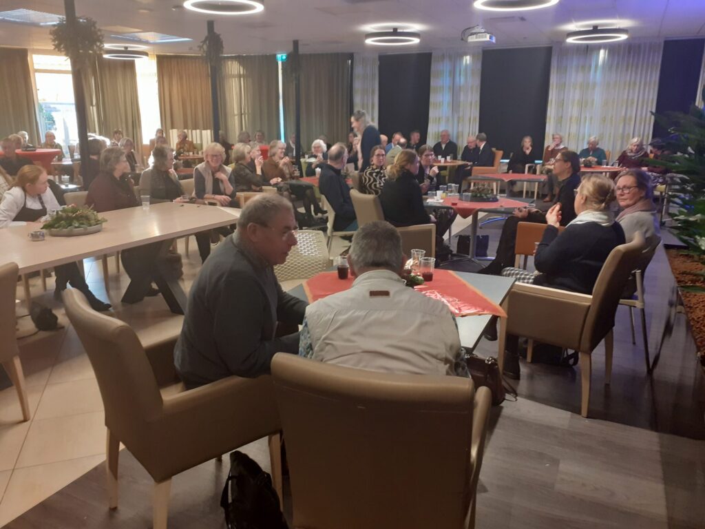 Een groep mensen zit in een zaal aan tafels, in gesprek tijdens een bijeenkomst. De ruimte is sfeervol verlicht en gezellig ingericht.