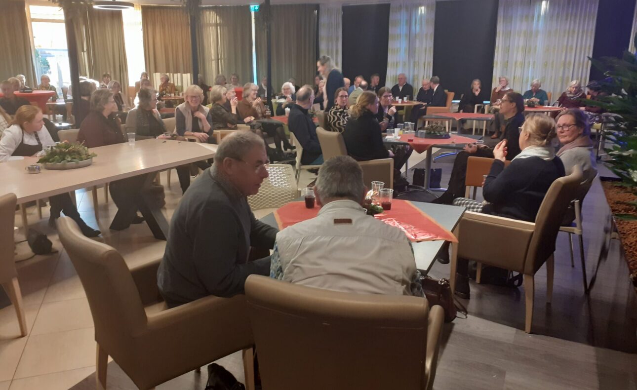 Een groep mensen zit in een zaal aan tafels, in gesprek tijdens een bijeenkomst. De ruimte is sfeervol verlicht en gezellig ingericht.