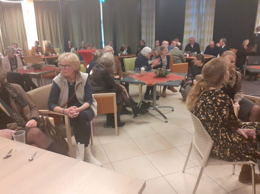 Groep mensen zit aan tafels in een zaal met gordijnen. Ze praten en lijken deel te nemen aan een sociale bijeenkomst.