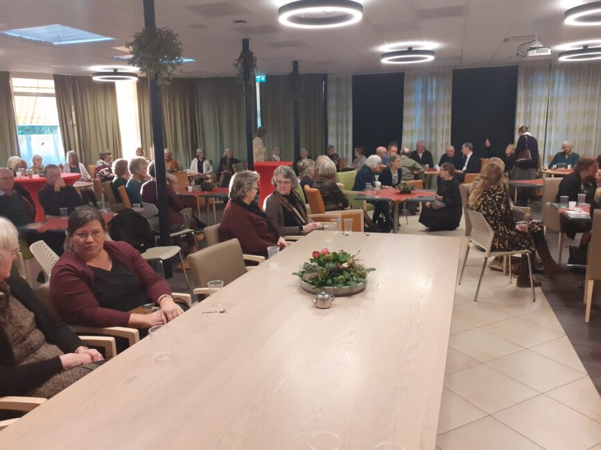 Een groep mensen zit in een gemeenschappelijke ruimte met tafels en stoelen. Er hangt een gezellige sfeer met kerstdecoratie.
