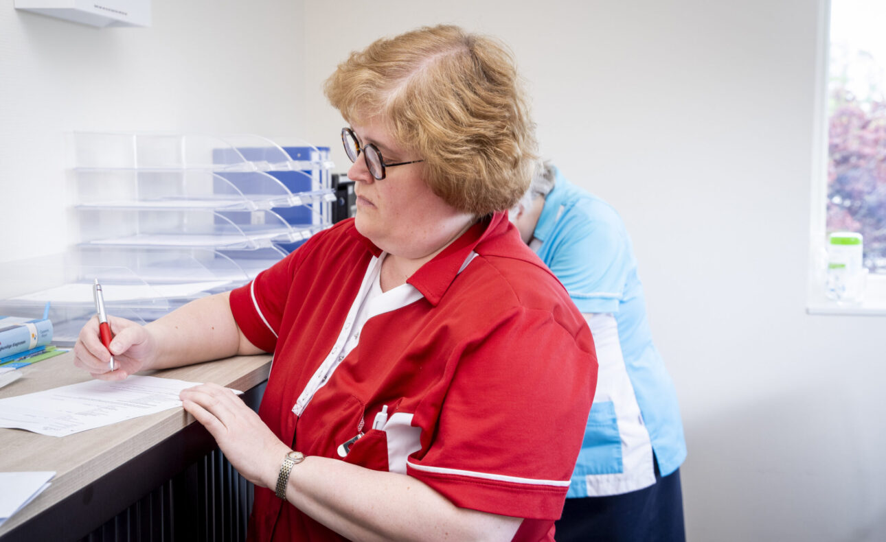 Een zorgmedewerker in een rood uniform schrijft iets op papier in een kantooromgeving met een collega op de achtergrond.