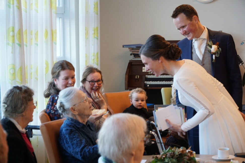 Een bruidspaar in gesprek met een oudere vrouw in een rolstoel, omringd door familieleden in een gezellige kamer.