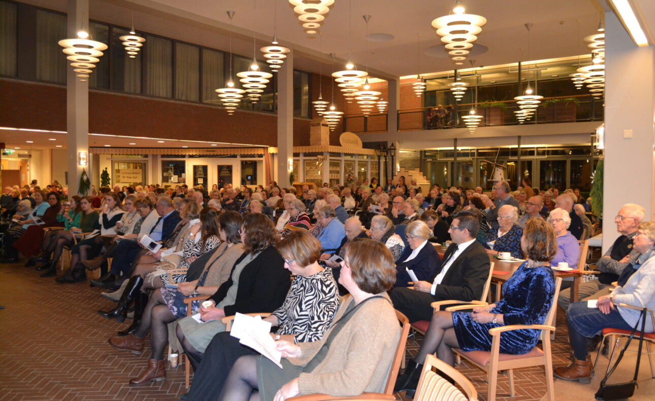Een grote groep mensen zit in een zaal met moderne verlichting, aandachtig luisterend naar een presentatie of spreker.