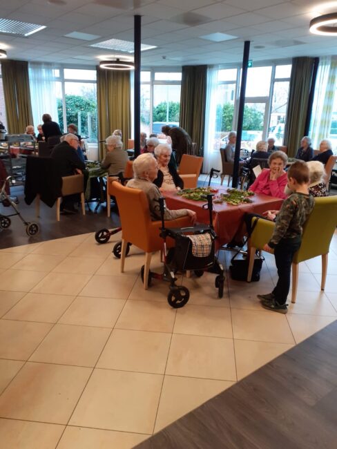 Groep ouderen zit aan tafel in een gemeenschappelijke ruimte, pratend en genietend van elkaars gezelschap. Een kind staat erbij.
